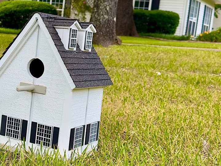 White cottage birdhouse with dormer windows on the lawn in front of the matching home
